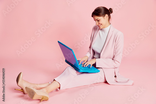 Portrait of beautiful young girl in suit and glasses posing, working on laptop over pink background. Business communication