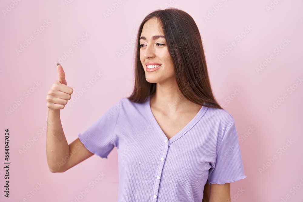 Young hispanic woman with long hair standing over pink background looking proud, smiling doing thumbs up gesture to the side Young hispanic woman with long hair standing over pink background looking proud, smiling doing thumbs up gesture to the side