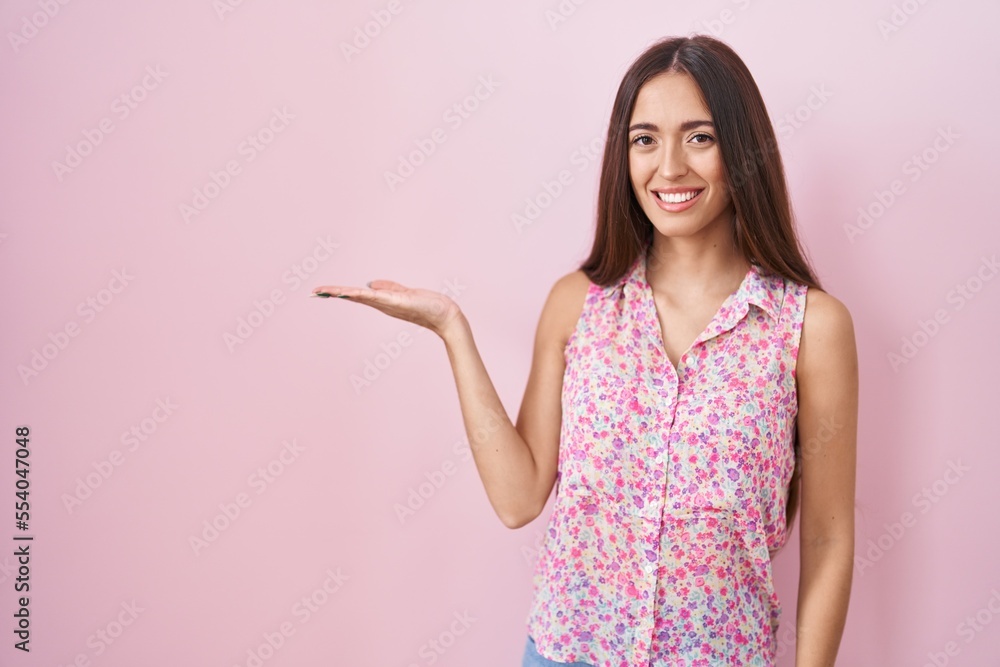 Young hispanic woman with long hair standing over pink background smiling cheerful presenting and pointing with palm of hand looking at the camera. Young hispanic woman with long hair standing over pink background smiling cheerful presenting and pointing with palm of hand looking at the camera.