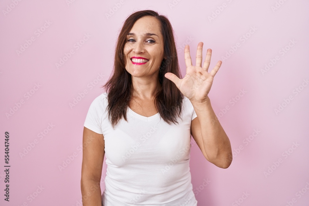 Middle age brunette woman standing over pink background showing and pointing up with fingers number five while smiling confident and happy.