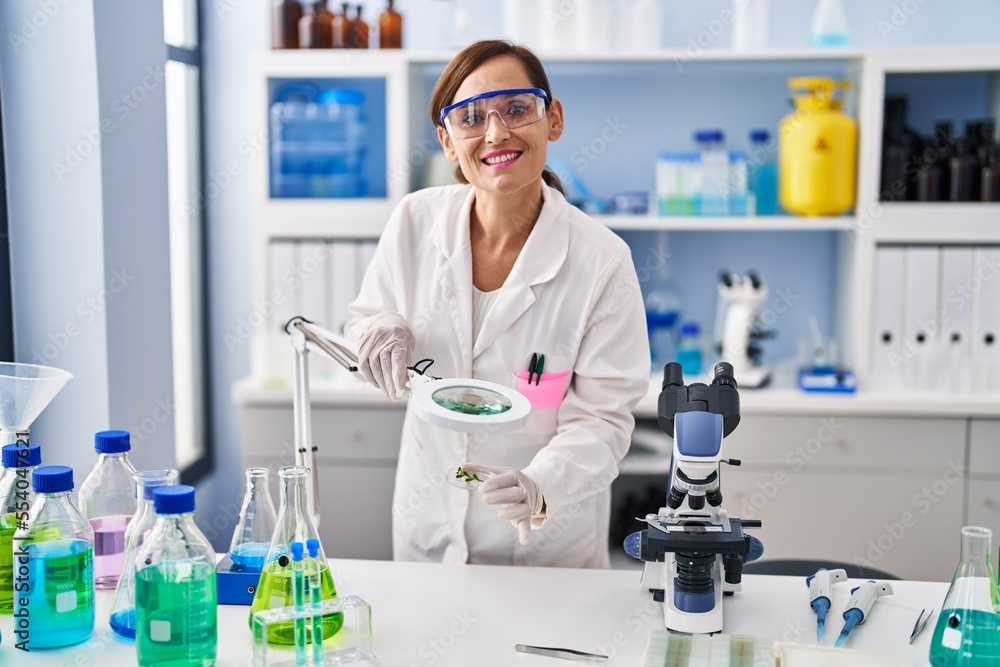 Middle age woman wearing scientist uniform looking herb with loupe at laboratory Middle age woman wearing scientist uniform looking herb with loupe at laboratory