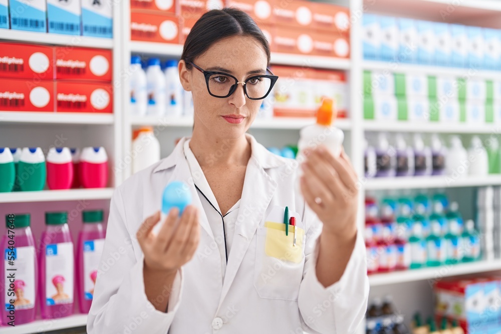 Young beautiful hispanic woman pharmacist holding deodorant bottles at pharmacy
