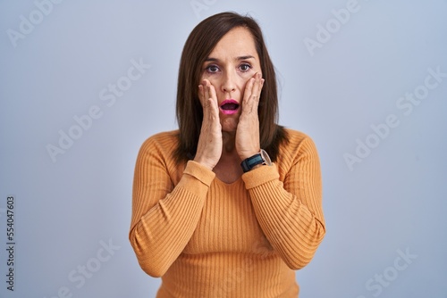 Middle age brunette woman standing wearing orange sweater afraid and shocked, surprise and amazed expression with hands on face