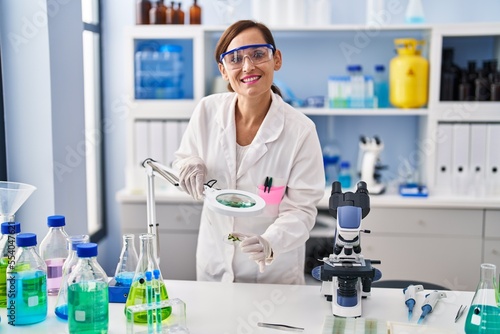 Middle age woman wearing scientist uniform looking herb with loupe at laboratory