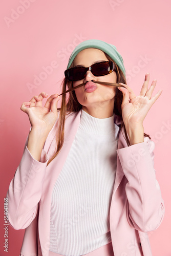 Portrait of young beautiful girl in a suit, hat and sunglasses posing over pink background. Good mood