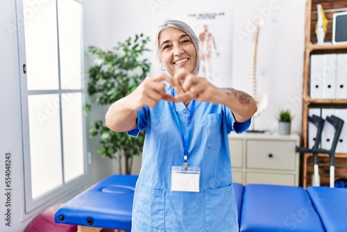 Middle age grey-haired woman wearing physiotherapist uniform at medical clinic smiling in love doing heart symbol shape with hands. romantic concept.