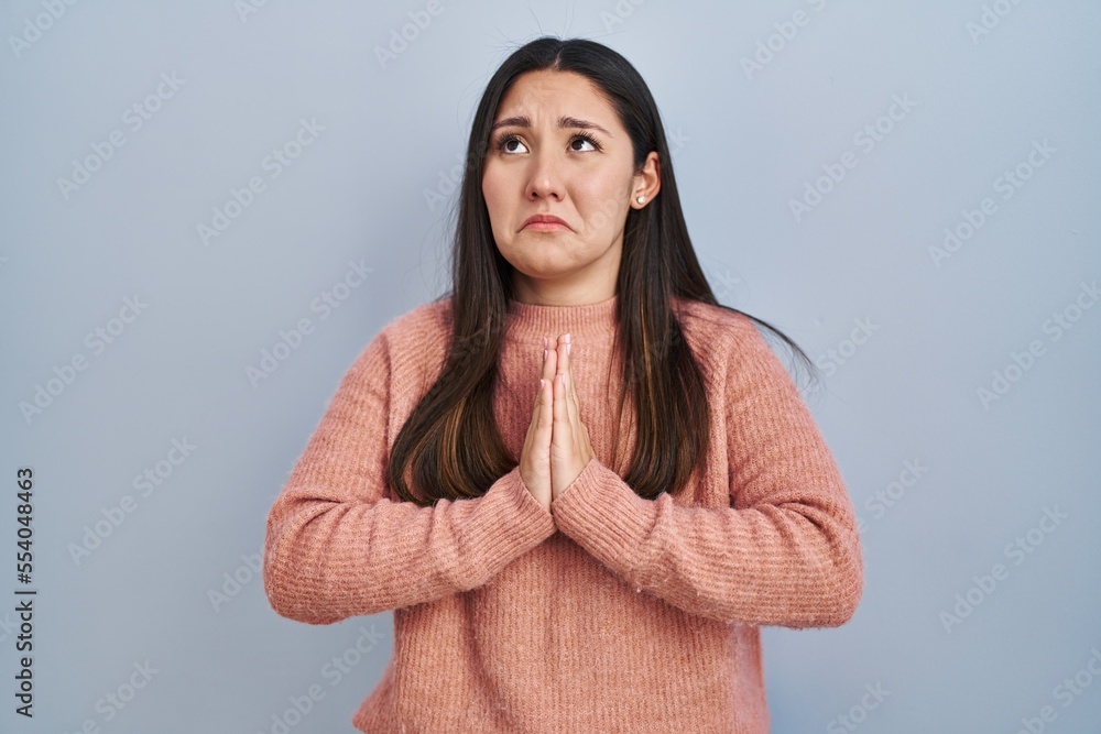 Fototapeta premium Young latin woman standing over blue background begging and praying with hands together with hope expression on face very emotional and worried. begging.