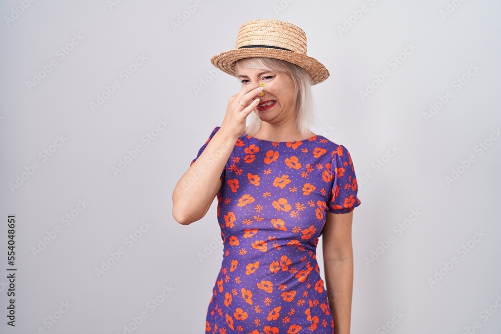 Young caucasian woman wearing flowers dress and summer hat smelling something stinky and disgusting, intolerable smell, holding breath with fingers on nose. bad smell Young caucasian woman wearing flowers dress and summer hat smelling something stinky and disgusting, intolerable smell, holding breath with fingers on nose. bad smell