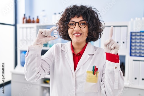 Hispanic doctor woman with curly hair holding vaccine surprised with an idea or question pointing finger with happy face  number one