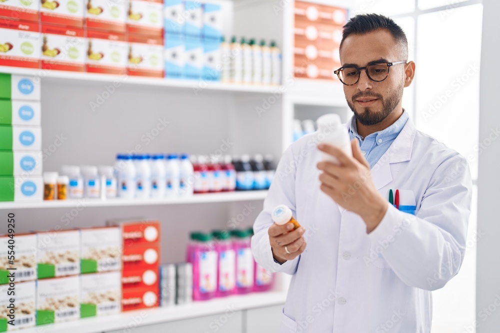 Young hispanic man pharmacist holding pills bottles at pharmacy