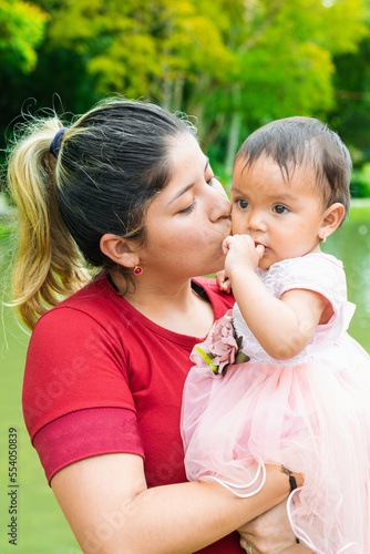 latina mother kissing her little brunette baby while holding her in her arms, baby distracted while being kissed on her cheek