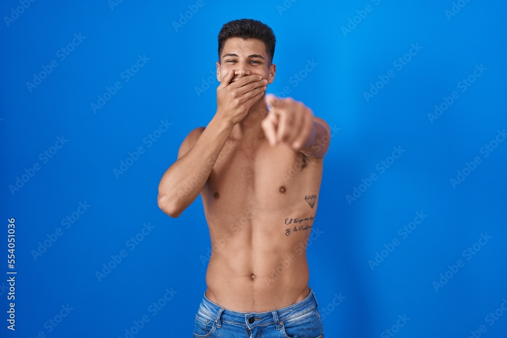 Young hispanic man standing shirtless over blue background laughing at you, pointing finger to the camera with hand over mouth, shame expression