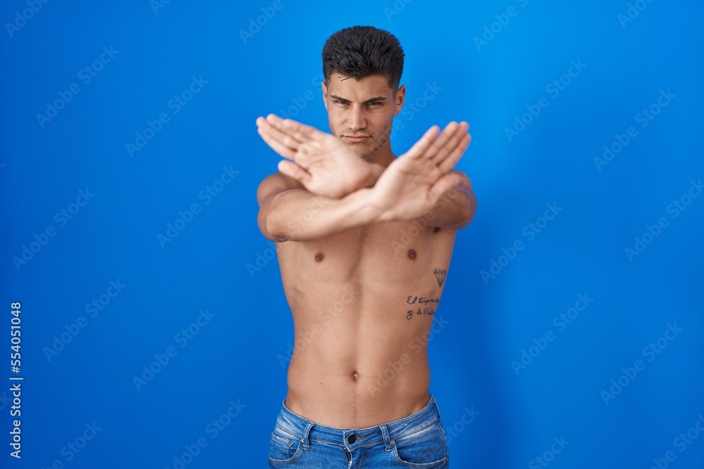 Young hispanic man standing shirtless over blue background rejection expression crossing arms and palms doing negative sign, angry face