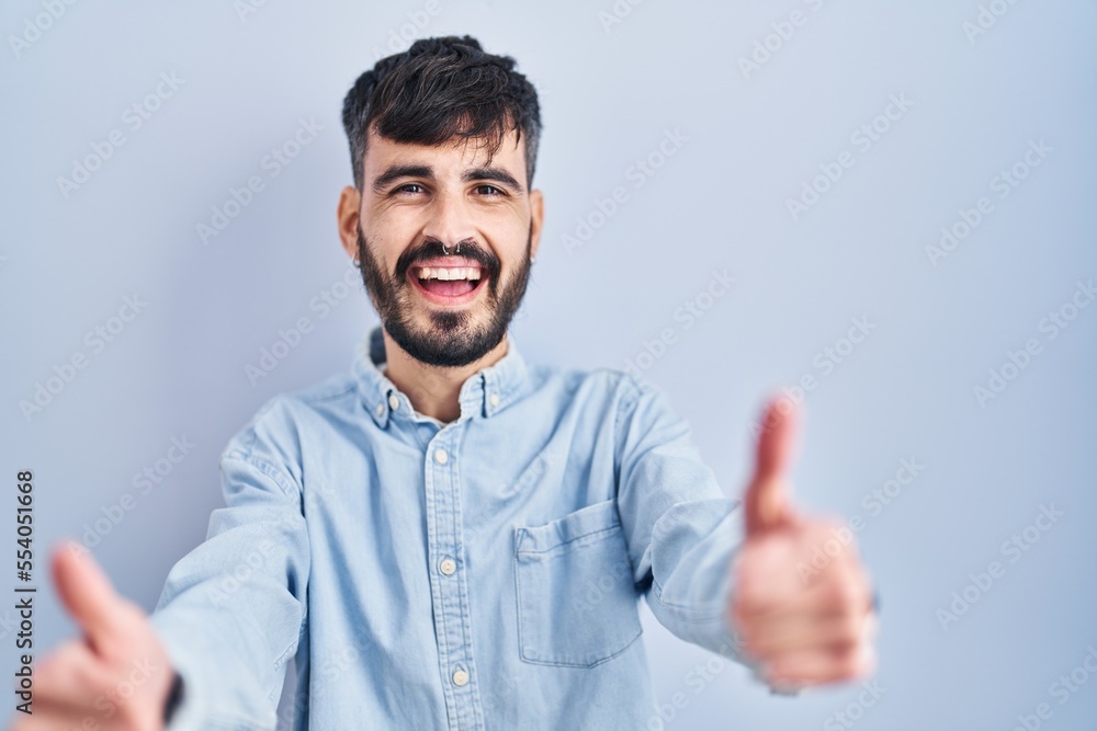 Young hispanic man with beard standing over blue background approving doing positive gesture with hand, thumbs up smiling and happy for success. winner gesture. Young hispanic man with beard standing over blue background approving doing positive gesture with hand, thumbs up smiling and happy for success. winner gesture.