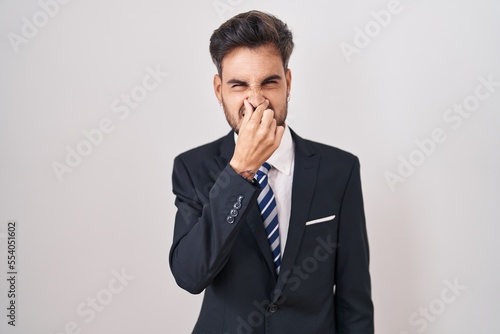 Young hispanic man with tattoos wearing business suit and tie smelling something stinky and disgusting, intolerable smell, holding breath with fingers on nose. bad smell