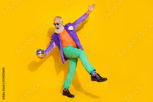 Full length photo of funky funny man wear purple velvet jacket holding discotheque ball having fun walking isolated yellow color background