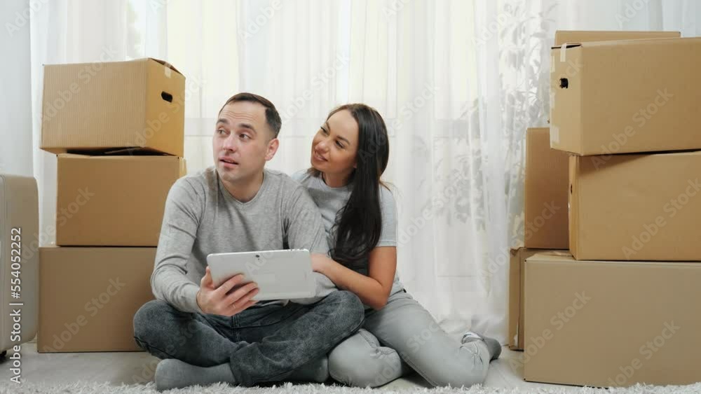 Man and woman sit in new apartment and look at tablet choosing decoration. Excited and amused married couple enjoys moving in new flat and sits among carton boxes Man and woman sit in new apartment and look at tablet choosing decoration. Excited and amused married couple enjoys moving in new flat and sits among carton boxes