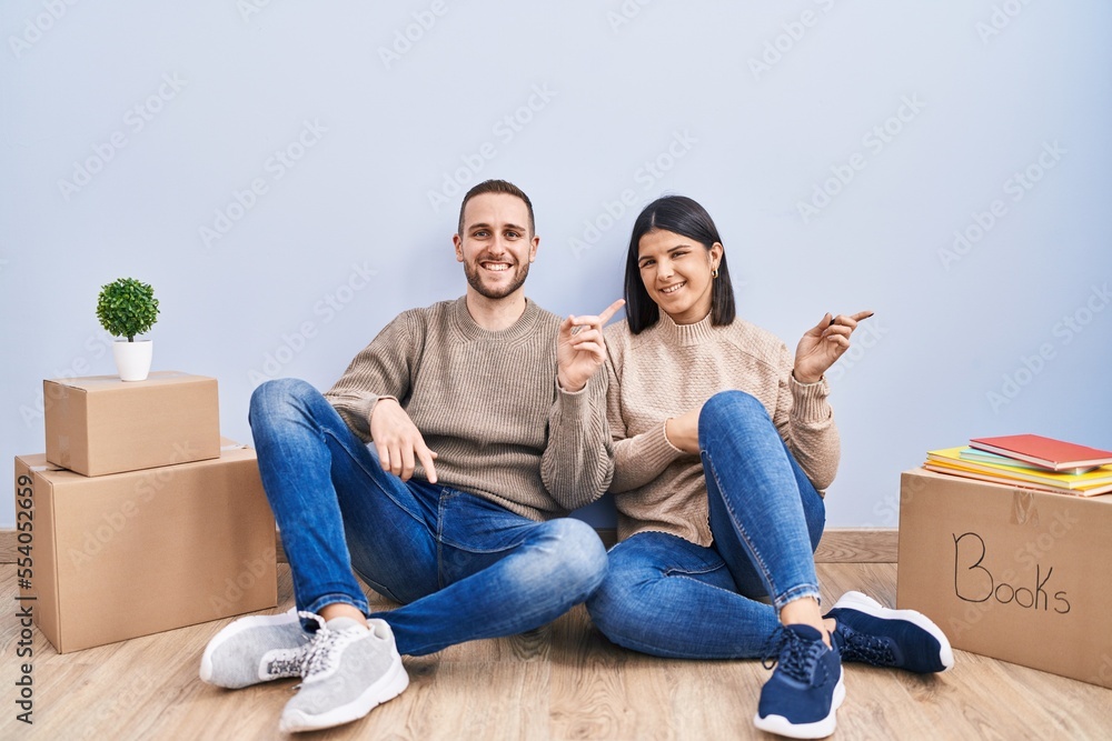 Young couple moving to a new home with a big smile on face, pointing with hand finger to the side looking at the camera.