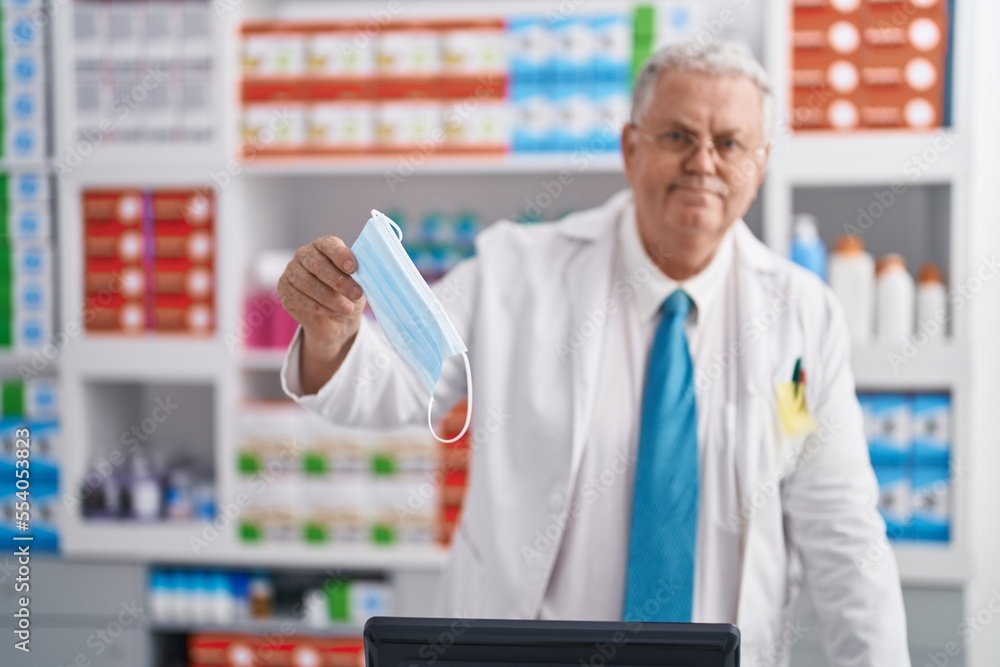 Middle age grey-haired man pharmacist smiling confident holding medical mask at pharmacy