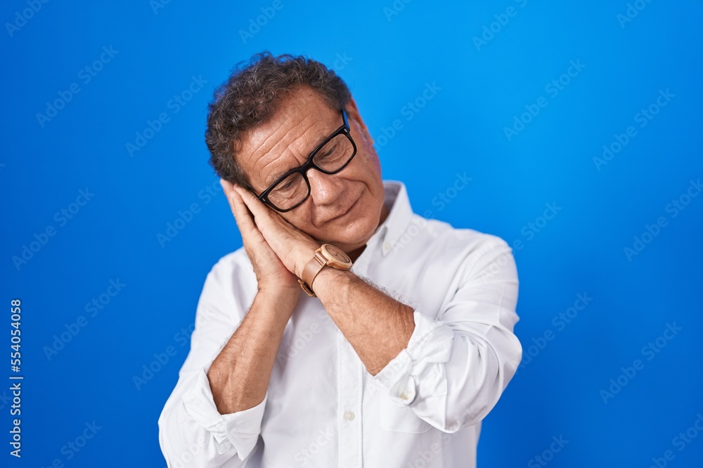 Middle age hispanic man standing over blue background sleeping tired dreaming and posing with hands together while smiling with closed eyes. Middle age hispanic man standing over blue background sleeping tired dreaming and posing with hands together while smiling with closed eyes.