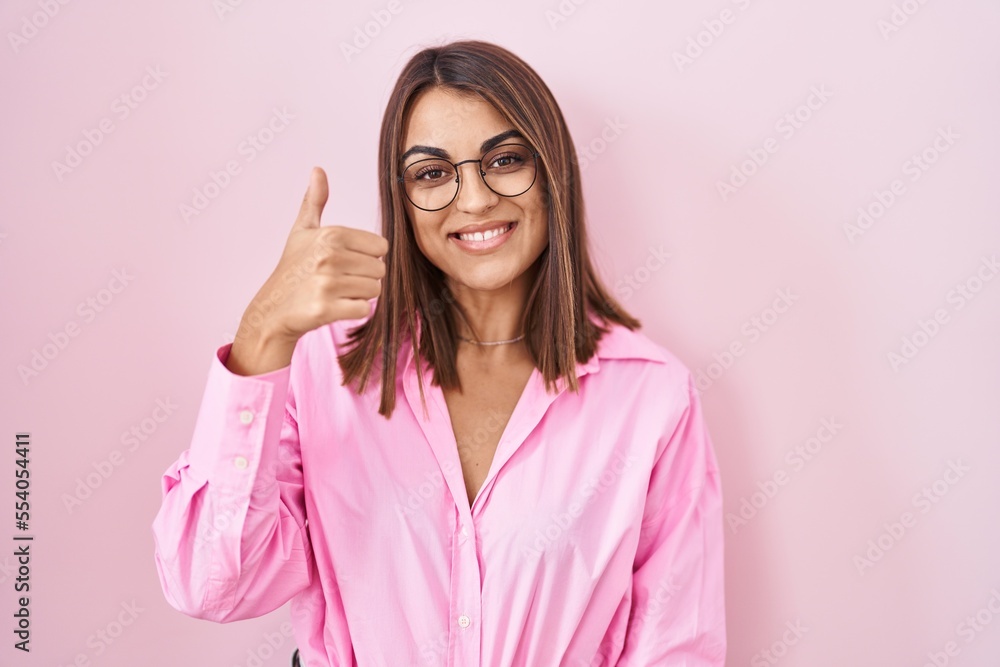 Young hispanic woman wearing glasses standing over pink background doing happy thumbs up gesture with hand. approving expression looking at the camera showing success. Young hispanic woman wearing glasses standing over pink background doing happy thumbs up gesture with hand. approving expression looking at the camera showing success.