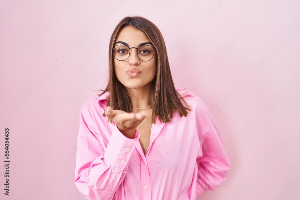 Young hispanic woman wearing glasses standing over pink background looking at the camera blowing a kiss with hand on air being lovely and sexy. love expression. Young hispanic woman wearing glasses standing over pink background looking at the camera blowing a kiss with hand on air being lovely and sexy. love expression.