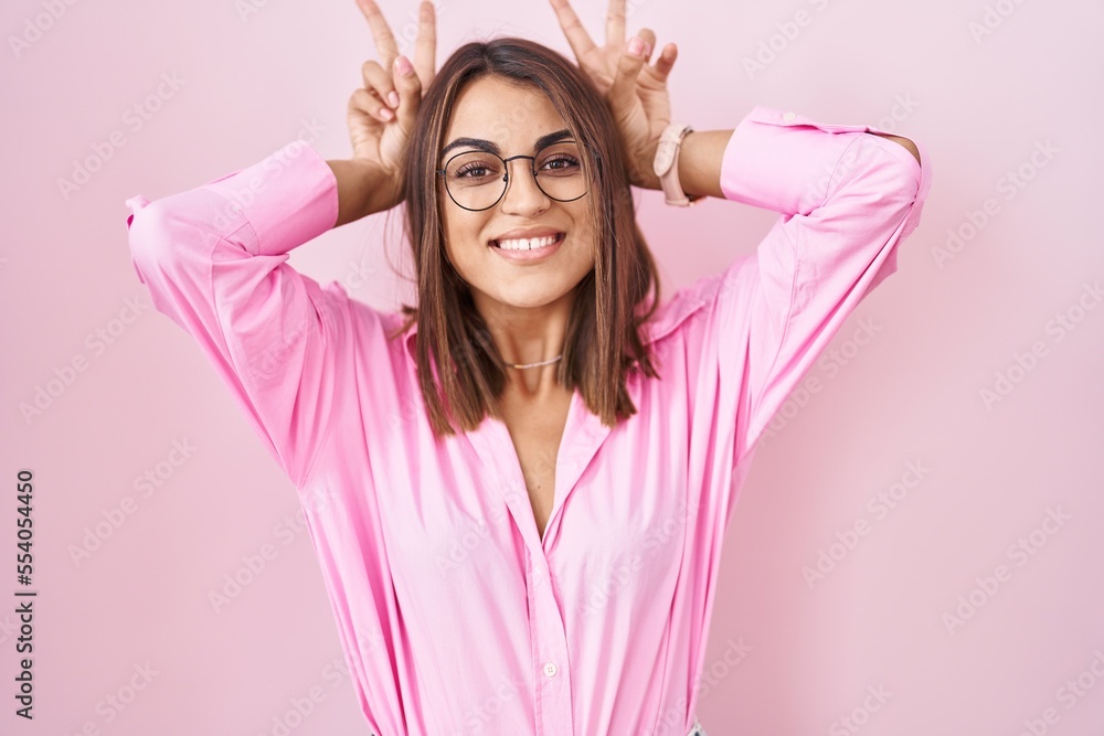 Young hispanic woman wearing glasses standing over pink background posing funny and crazy with fingers on head as bunny ears, smiling cheerful Young hispanic woman wearing glasses standing over pink background posing funny and crazy with fingers on head as bunny ears, smiling cheerful