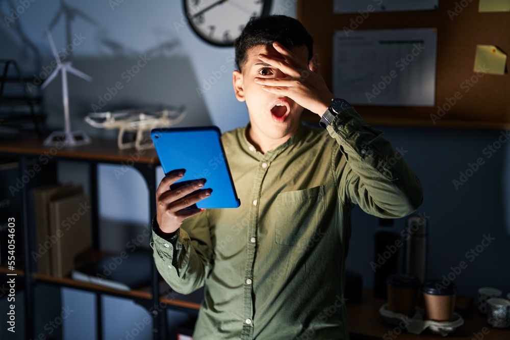 Non binary person using touchpad device at night peeking in shock covering face and eyes with hand, looking through fingers with embarrassed expression. Non binary person using touchpad device at night peeking in shock covering face and eyes with hand, looking through fingers with embarrassed expression.
