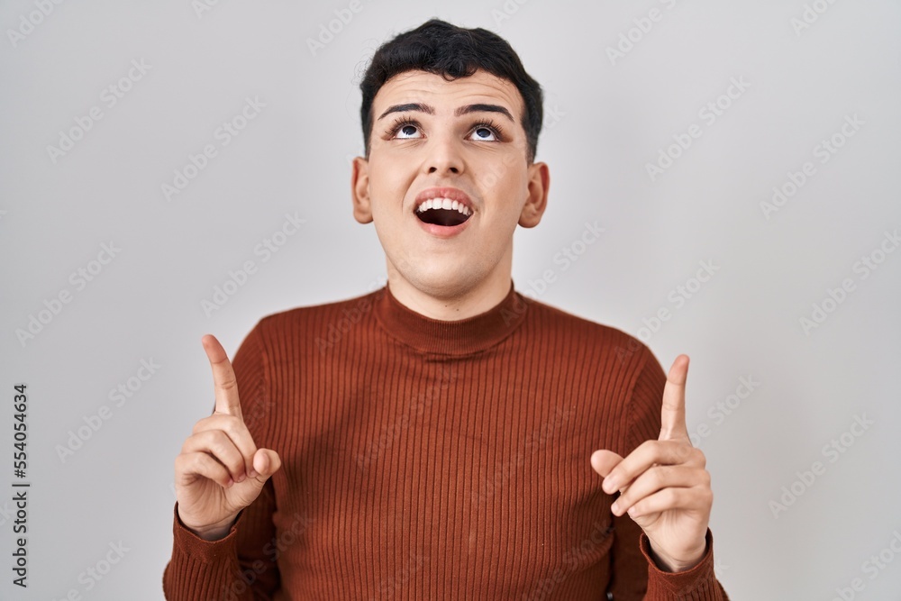 Non binary person wearing make up standing over isolated background amazed and surprised looking up and pointing with fingers and raised arms. Non binary person wearing make up standing over isolated background amazed and surprised looking up and pointing with fingers and raised arms.