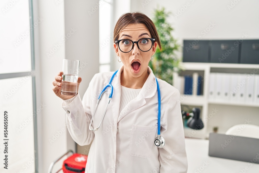 Φωτογραφία Young brunette doctor woman holding glass of water scared and amazed with open m