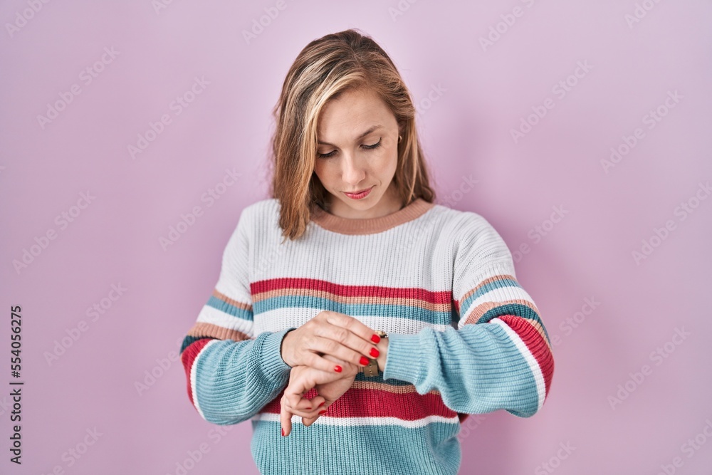 Young blonde woman standing over pink background checking the time on wrist watch, relaxed and confident Young blonde woman standing over pink background checking the time on wrist watch, relaxed and confident