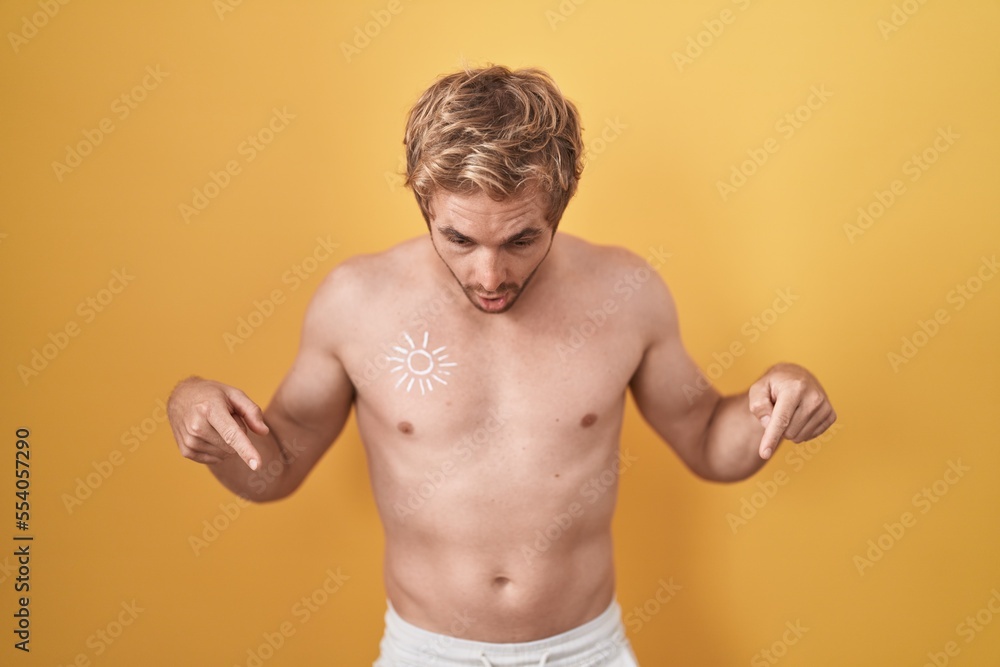 Caucasian man standing shirtless wearing sun screen pointing down with fingers showing advertisement, surprised face and open mouth