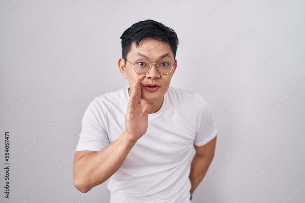 Fototapeta premium Young asian man standing over white background hand on mouth telling secret rumor, whispering malicious talk conversation
