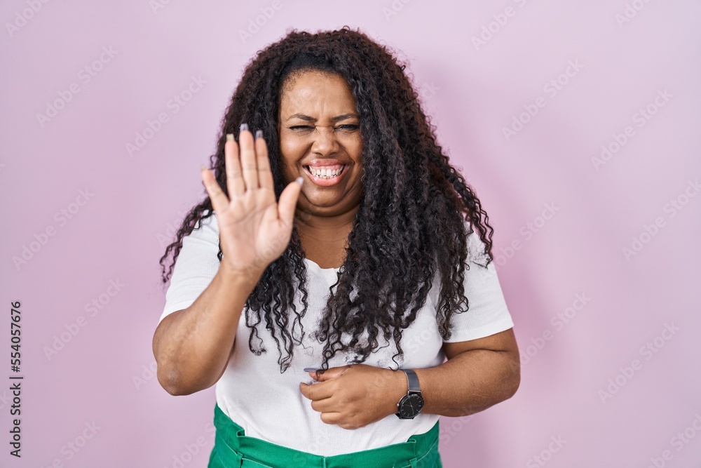 Plus size hispanic woman standing over pink background waiving saying hello happy and smiling, friendly welcome gesture Plus size hispanic woman standing over pink background waiving saying hello happy and smiling, friendly welcome gesture