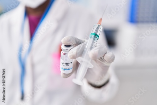 African american woman wearing scientist uniform and medical mask holding covid-19 vaccine at laboratory