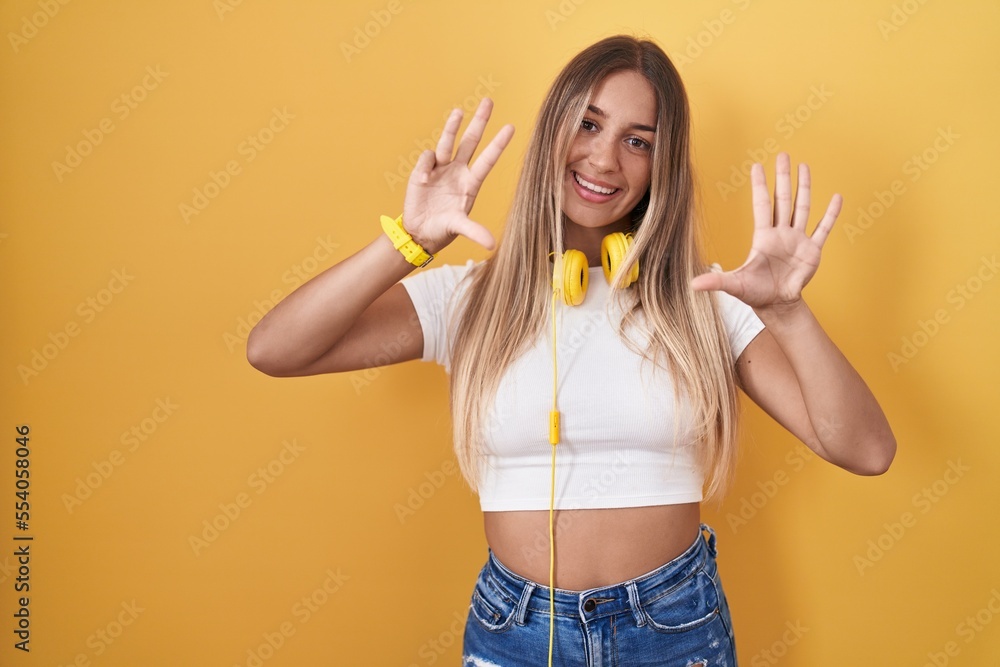 Young blonde woman standing over yellow background wearing headphones showing and pointing up with fingers number nine while smiling confident and happy. Young blonde woman standing over yellow background wearing headphones showing and pointing up with fingers number nine while smiling confident and happy.