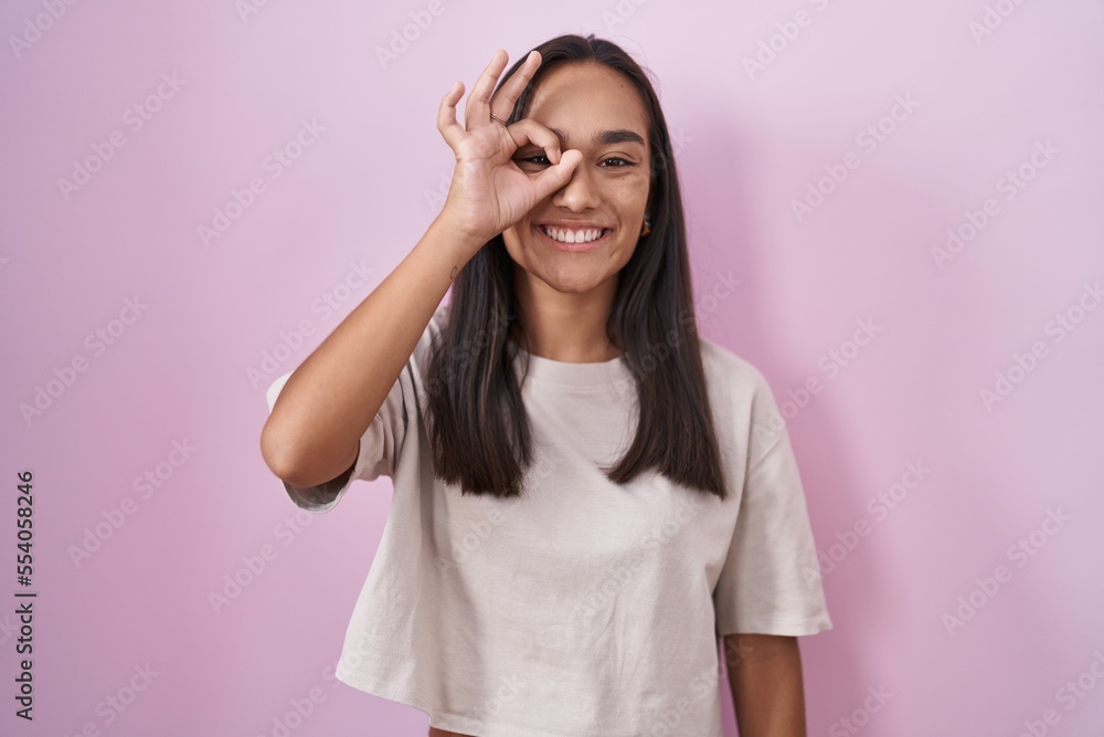 Young hispanic woman standing over pink background doing ok gesture with hand smiling, eye looking through fingers with happy face. Young hispanic woman standing over pink background doing ok gesture with hand smiling, eye looking through fingers with happy face.
