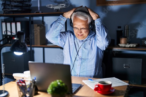 Hispanic senior man wearing call center agent headset at night suffering from headache desperate and stressed because pain and migraine. hands on head.