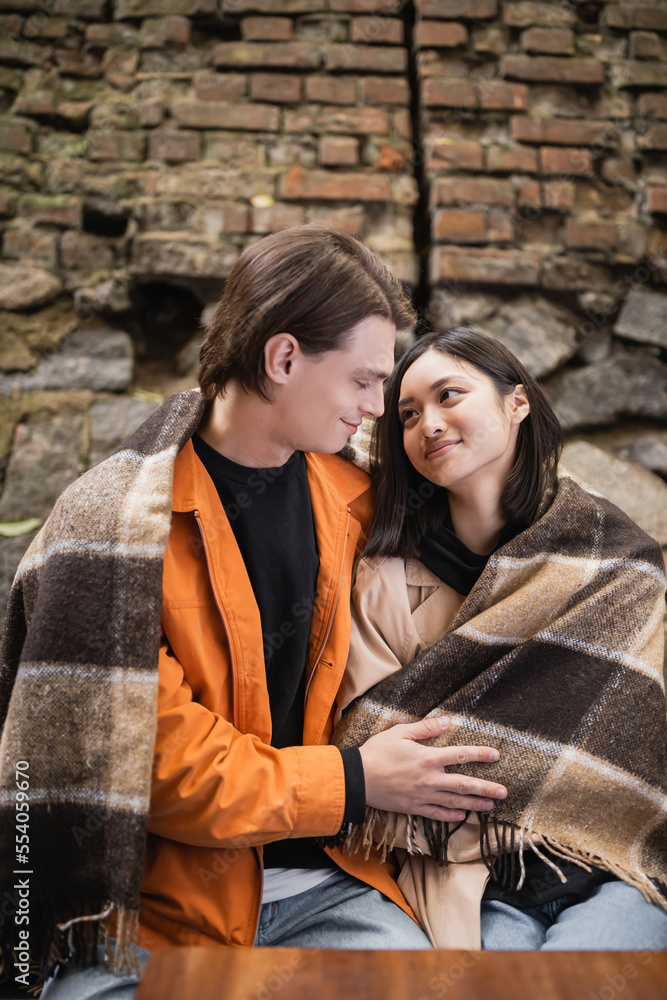 Smiling interracial couple in blanket looking at each other while sitting in blanket in outdoor cafe.