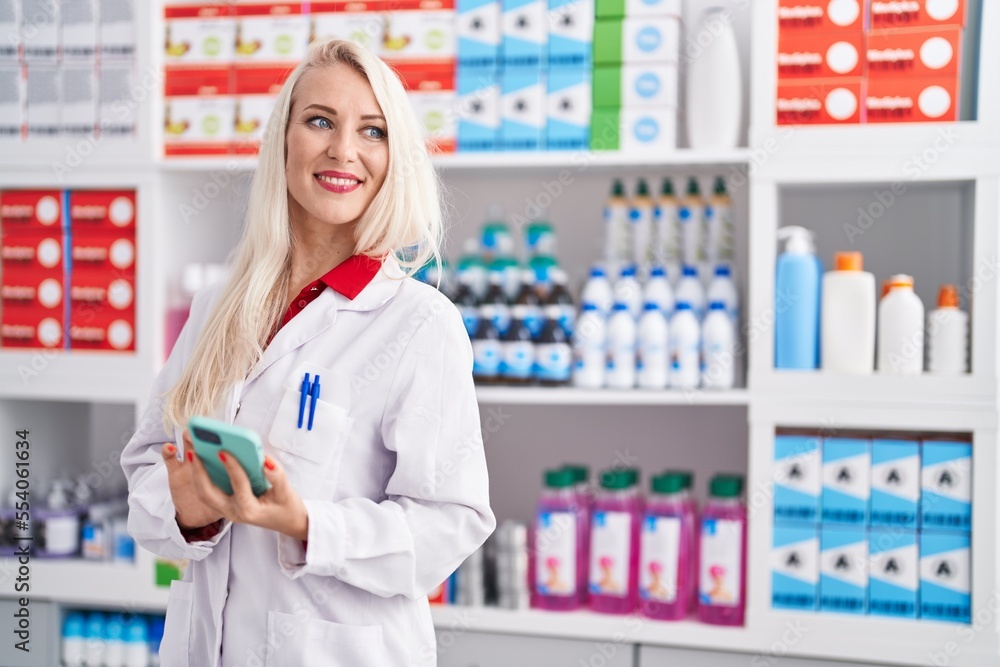 Young blonde woman pharmacist using smartphone working at pharmacy
