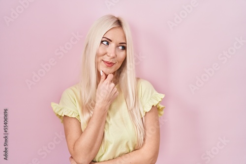 Caucasian woman standing over pink background with hand on chin thinking about question, pensive expression. smiling and thoughtful face. doubt concept.
