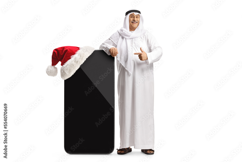 Mature arab man in traditional clothes leaning on a big mobile phone with a santa hat and pointing