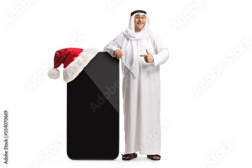 Mature arab man in traditional clothes leaning on a big mobile phone with a santa hat and pointing