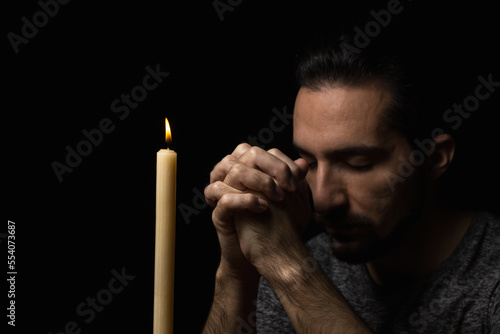 A man folded his hands in prayer next to a burning candle on a black background. prayer to God for happiness and a better life. Repent of your sins. Unity with God