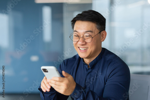 Cheerful and smiling asian businessman inside office using phone, man typing message and browsing online pages and watching video, boss in glasses and shirt at work.