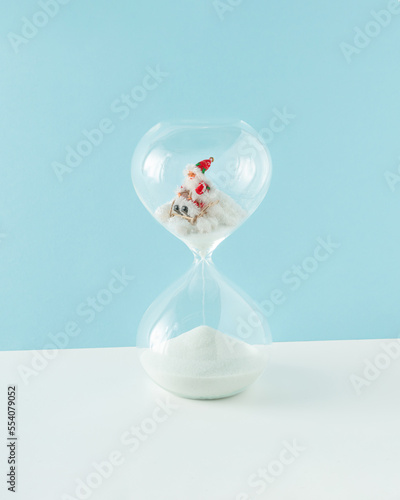 2023. Santa Claus on his sleigh makes his way through the snow in the hourglass on isolated pastel blue and white background. Minimal New Years greeting card. Xmas time or waiting santa concept.