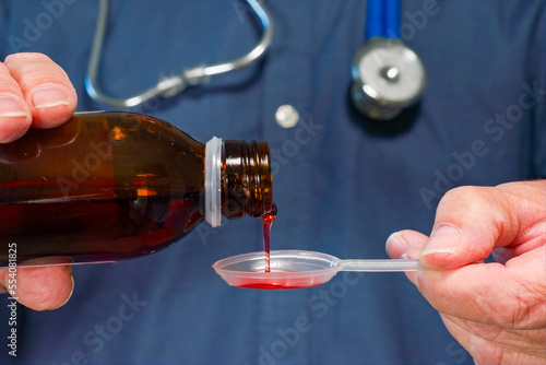 A doctor pours medicine from a bottle into a plastic spoon prior to administering it to a patient. Selective focus on the stream of medicine going into the spoon. Hospital themed medical image 