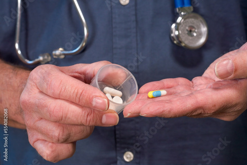 A doctor places medical pills and tablets into a plastic pot prior to administering them to a patient. Selective focus on the blue tablet in the doctors hand. Hospital theme medical image. Stethoscope