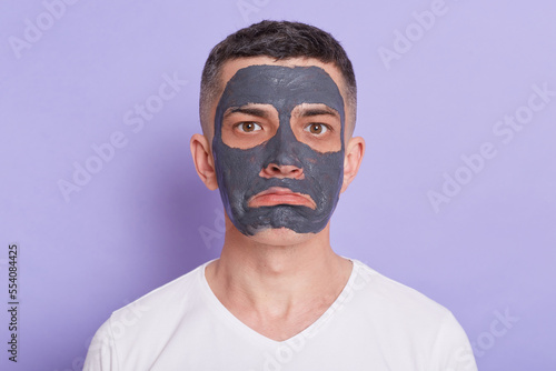 Portrait of handsome brunet man with sad expression, wears nourishing homemade mud mask for reducing dark dotes, wears casual white t shirt, isolated over purple background.