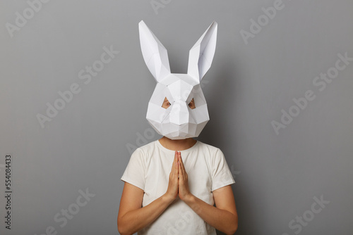 Portrait of unknown woman wearing white t shirt and paper rabbit mask standing isolated over gray background, keeping hands in prayer gesture, practicing yoga, meditating. photo
